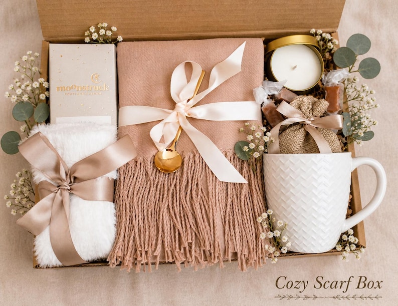 May include: A gift box containing a dusty rose fringed scarf tied with a cream ribbon and gold spoon, a white faux fur item with a satin ribbon, a white textured mug, a candle in a gold tin, a small burlap bag, and a box labeled moonstruck.