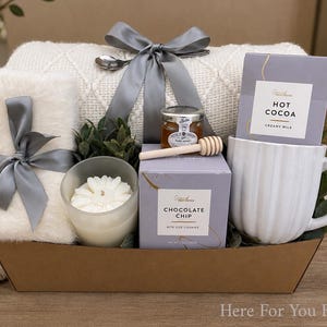 May include: A gift basket containing a white textured throw blanket tied with a grey ribbon, a white faux fur item with a grey ribbon, a white candle, a jar of honey with a wooden dipper, a box of chocolate chip cookies, and a white ribbed mug with hot cocoa.