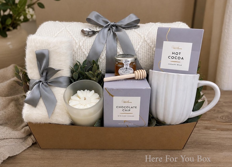 May include: A gift basket containing a white textured throw blanket tied with a grey ribbon, a white faux fur item with a grey ribbon, a white candle, a jar of honey with a wooden dipper, a box of chocolate chip cookies, and a white ribbed mug with hot cocoa.