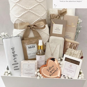 May include: A white gift box filled with self-care items, including a blanket, tumbler, chocolates, and a heart-shaped dish with the text "Follow your dreams!". The box is decorated with flowers and a ribbon.