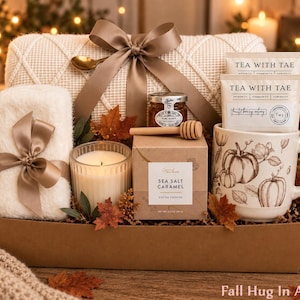 May include: A gift box containing a cream-colored knit blanket with a brown ribbon, a white fuzzy cloth, a glass candle, a jar of honey with a wooden dipper, a box of Sea Salt Caramel cookies, and a mug with pumpkin illustrations and tea bags.