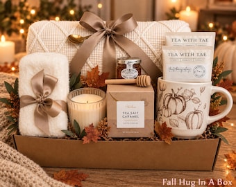 May include: A gift box containing a cream-colored knit blanket with a brown ribbon, a white fuzzy cloth, a glass candle, a jar of honey with a wooden dipper, a box of Sea Salt Caramel cookies, and a mug with pumpkin illustrations and tea bags.