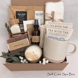 May include: A gift box containing a speckled ceramic mug with two Tea With Tae packets, a candle with flower-shaped wax, a glass jar of bath salts, a bar of oatmeal soap, a bottle of oat lotion, two Gel-Ohh! packets, a white fuzzy item, and a loofah.