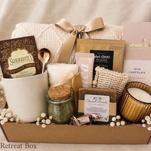 May include: A gift box containing a white ceramic mug, a packet of Sorrento chocolate truffle hot cocoa mix, a gold spoon, a cream-colored knit blanket with a satin bow, a jar of bath salts, a bar of soap, a candle, and Moonstruck milk chocolate.