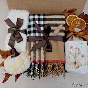 May include: A gift box containing a white fuzzy item, a tan and black plaid scarf with fringe, a white ceramic mug with pumpkin illustrations, a small white pumpkin, dried orange slices, a brown ribbon, and a rose gold spoon, labeled Cozy Fall Box.