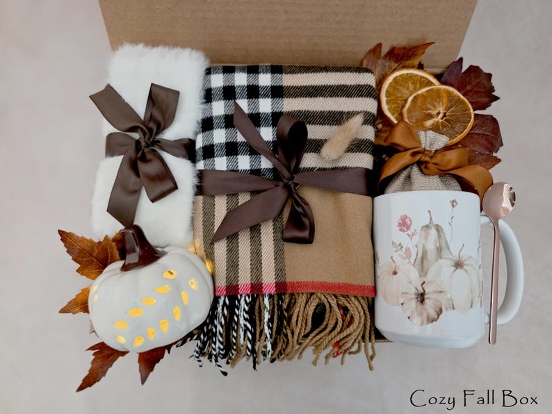 May include: A gift box containing a white fuzzy item, a tan and black plaid scarf with fringe, a white ceramic mug with pumpkin illustrations, a small white pumpkin, dried orange slices, a brown ribbon, and a rose gold spoon, labeled Cozy Fall Box.