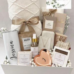 May include: A white gift box filled with self-care items, including a blanket, tumbler with "Jessica" on it, chocolates, and bath products. A "Happy Birthday" card and a heart-shaped dish are also included. The box is decorated with flowers and a ribbon.