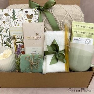 May include: A gift box containing a notebook, chocolate, tea, a mug, a candle, and a small blanket. The box is tied with a green ribbon. The items are arranged in a brown box, creating a cosy and inviting aesthetic. The box is labelled "Green Floral Box".