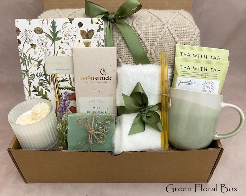 May include: A gift box containing a notebook, chocolate, tea, a mug, a candle, and a small blanket. The box is tied with a green ribbon. The items are arranged in a brown box, creating a cosy and inviting aesthetic. The box is labelled "Green Floral Box".