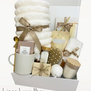 May include: A white gift box containing self-care products. It features a stack of white fluffy towels tied with a gold ribbon, a mug with hot cocoa, a candle, bath products, and a wooden handled brush. The box is labelled "Linen Luxe Box".