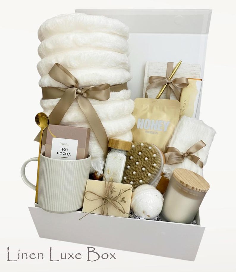 May include: A white gift box containing self-care products. It features a stack of white fluffy towels tied with a gold ribbon, a mug with hot cocoa, a candle, bath products, and a wooden handled brush. The box is labelled "Linen Luxe Box".