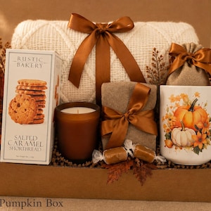 May include: A gift box containing a white knit blanket with a brown ribbon, a box of Rustic Bakery Salted Caramel Shortbread, a brown candle, a wrapped gift, a burlap pouch, a white mug with pumpkin graphics, a gold spoon, and autumn leaves.