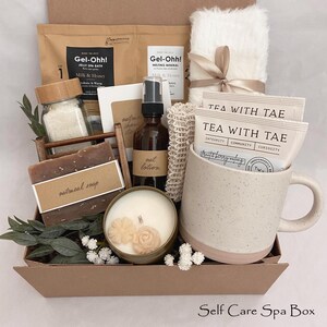 May include: A self-care spa box containing a selection of items. It features a mug, tea bags, a candle, soap, bath products, and a fluffy white towel tied with a ribbon. The box is brown with the text "Self Care Spa Box".
