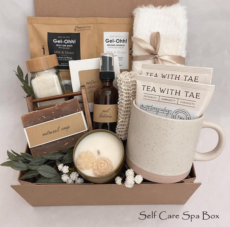 May include: A self-care spa box containing a selection of items. It features a mug, tea bags, a candle, soap, bath products, and a fluffy white towel tied with a ribbon. The box is brown with the text "Self Care Spa Box".