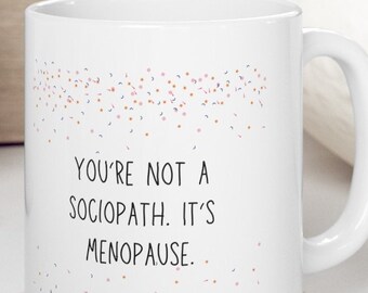 May include: White ceramic coffee mug with a black and white text design that reads "You're not a sociopath. It's menopause." The mug has a white handle and is decorated with a light pink and blue confetti pattern.