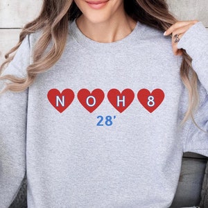Op de afbeelding: Grijs sweatshirt met rode harten en de tekst "NOH8" en "28'" in blauw.