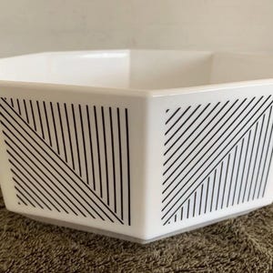 May include: A white hexagonal bowl featuring a black geometric pattern of diagonal lines on each side. The bowl has a smooth, glossy finish and a wide, open top, showcasing a clean, angular design with sharp edges and a consistent, repeating motif.