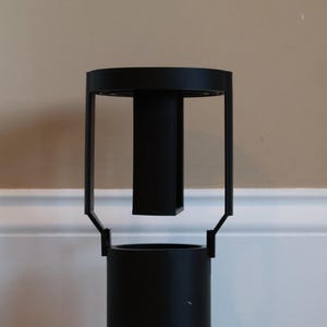 Op de afbeelding: Een zwarte metalen decoratieve standaard met een cilindrische basis en een rechthoekige bovenkant. De standaard heeft een modern ontwerp met een verticale steun en een cirkelvormige bovenkant. De standaard is ongeveer 30 cm hoog.