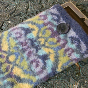 May include: A blue, yellow and purple knitted tablet sleeve with a black button closure. The sleeve is made of a soft, fuzzy material and has a geometric pattern.