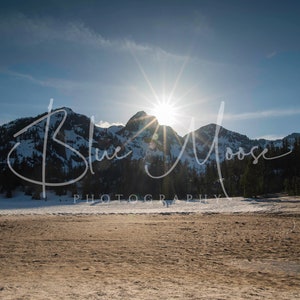 Könnte beinhalten: Landschaftsfotografie mit schneebedeckten Bergen unter einem hellen Sonnenaufgang. Das Bild, mit der Marke "Blue Moose Photography", zeigt eine ruhige Szene im Freien mit einem Vordergrund aus trockenem, hellbraunem Gelände.