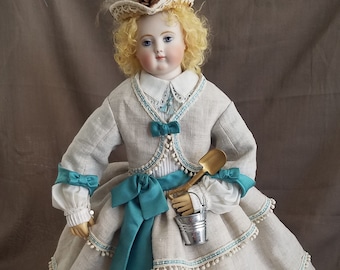 May include: A porcelain doll wearing a white dress with a teal sash and a white jacket. The doll is holding a small bucket and shovel. The doll has blonde hair and blue eyes.