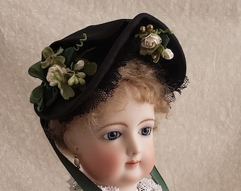 May include: A porcelain doll with blue eyes and blonde hair wearing a black hat with green and white flowers and a green ribbon. The doll is wearing a black dress with a white lace collar and a pearl necklace.