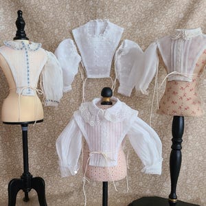 Può includere: Quattro camicette bianche per bambole con bordi in pizzo e maniche a sbuffo, esposte su manichini. Le camicette sono realizzate in un tessuto trasparente e hanno un aspetto delicato e femminile.