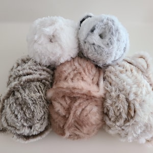 May include: Six balls of fluffy yarn in a range of neutral shades. The colours are white, grey, brown, and a pale pink. The yarn has a soft, textured look, indicating it's made from a plush material, suitable for knitting or crochet projects.