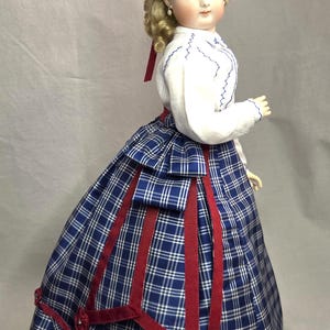 May include: A porcelain doll with blonde curly hair wearing a white long-sleeved blouse with blue stitching and a blue and white plaid skirt. The skirt features dark red ribbon trim and small red fabric rosettes. A matching blue plaid hat with red floral accents sits atop the head.