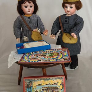 Puede incluir: Dos muñecas antiguas de porcelana con vestidos grises y sombreros verde oliva, con bolsos a juego. Un juego de mesa vintage está sobre una pequeña mesa de madera, y otra caja de juego está en el suelo. La caja del juego tiene el texto "NOUVEAU JEU DE SOCIETE".