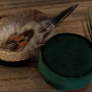 May include: Two decorative hats on a wooden surface. One hat is a woven design with feathers in shades of brown, tan, and black. The other is a dark green, round hat with a few green feathers.