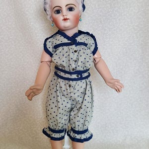 May include: A vintage porcelain doll with blue eyes, wearing a star-patterned romper with navy blue trim and matching bow. The doll has a white wig, turquoise earrings, and navy blue shoes. The doll is standing on a clear stand.