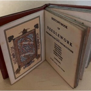 Op de afbeelding: Een open antiek boek met een rode kaft en vergeelde pagina's. De titelpagina luidt "Beeton's Book of Needlework" met instructies voor breien, haken en borduren. Het boek is opengeslagen op een decoratieve pagina.