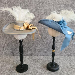 Può includere: Due cappelli su supporti neri. Il cappello a sinistra è grigio con un nastro dorato e una piuma bianca. Il cappello a destra è blu con una piuma bianca e un nastro blu.