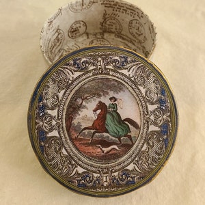 May include: A round, gold-trimmed trinket box with a blue and gold border. The lid features a colorful image of a woman riding a horse through a forest with a dog running behind her.