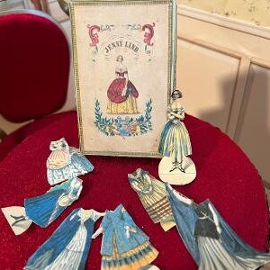 May include: A vintage paper doll set featuring Jenny Lind, a famous Swedish opera singer. The set includes a box with a portrait of Jenny Lind, a doll, and several paper outfits. The doll and outfits are on a red surface.