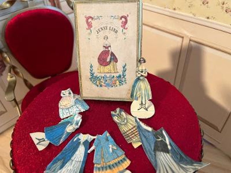 May include: A vintage paper doll set featuring Jenny Lind, a famous Swedish opera singer. The set includes a box with a portrait of Jenny Lind, a doll, and several paper outfits. The doll and outfits are on a red surface.