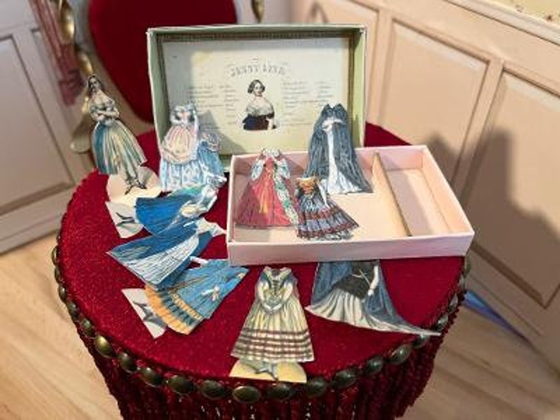 May include: A vintage paper doll set in a pink box with a red velvet base. The set includes a variety of paper dolls with different dresses and accessories. The box has the text "Jenny Lind" printed on the front.