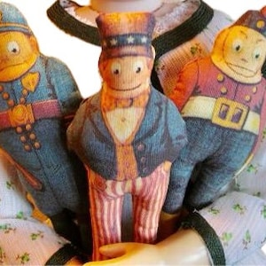 May include: Three vintage-style fabric dolls dressed in different outfits. The doll in the centre is wearing a blue and red striped suit with a top hat and a white bow tie. The doll on the left is wearing a blue police uniform with a yellow hat. The doll on the right is wearing a red and gold military uniform with a yellow hat.