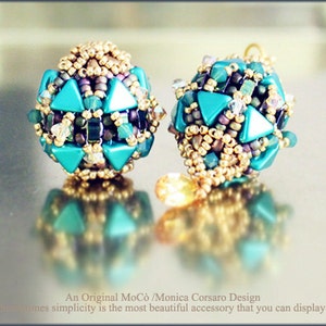 Puede incluir: Un par de pendientes hechos a mano con cuentas triangulares turquesas y cuentas de semillas doradas. Los pendientes tienen forma esférica y se reflejan en una superficie brillante. La imagen incluye el texto: "An Original MoCò /Monica Corsaro Design".