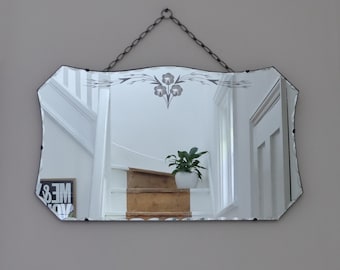 May include: A rectangular wall mirror with scalloped edges and a dark metal chain for hanging. The glass features a decorative etched floral design at the top center. The mirror reflects a staircase with wooden steps and a white railing.