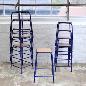 Può includere: Diversi sgabelli in metallo blu con sedili in legno chiaro sono impilati e disposti davanti a un muro di mattoni bianchi. Uno sgabello è da solo, mentre altri sono impilati in gruppi di tre e cinque. Design industriale semplice.
