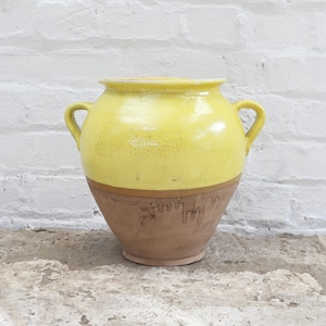Può includere: Un vaso in ceramica giallo e marrone con due manici. La metà superiore è di un giallo brillante, mentre la metà inferiore è marrone. Il vaso ha un'ampia apertura e due manici curvi. Probabilmente per scopi decorativi.