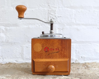 May include: A vintage wooden coffee grinder with a metal crank and a small drawer. The grinder has a light brown wooden body with the text "De Ve" and a logo on the front. The handle is made of wood.