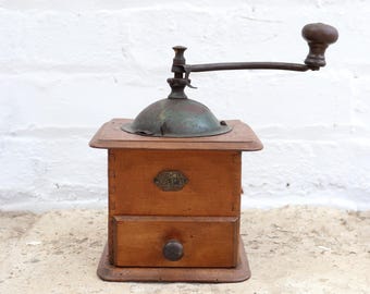 May include: A vintage wooden coffee grinder with a metal handle and a small drawer. The grinder has a square wooden base and a metal top. The handle is dark brown, and the metal top has a green patina. The drawer has a round wooden knob.