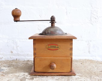 May include: A vintage wooden coffee grinder with a hand crank and a small drawer. The grinder has a metal top and a wooden handle. The label on the front reads "ODAX MADE IN FRANCE".