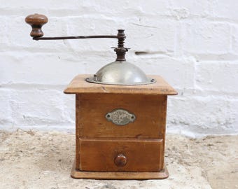 May include: A wooden coffee grinder with a square base, a small pull-out drawer with a round knob, and a metal dome top. A long metal handle with a wooden knob extends from the top. A metal plate on the front features the text Leinbrock's Ideal.