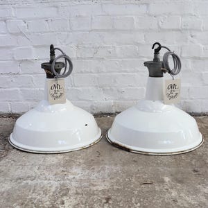 May include: Two white pendant lamps with a wide, bowl-shaped shade. Each lamp has a black and grey cord and a tag that reads "OWL & the elephant". The lamps are set against a white brick wall.