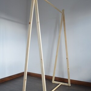 May include: A light-colored wooden clothing rack with an A-frame design. The rack has a horizontal bar for hanging clothes and is constructed from natural wood. The design is simple and minimalist, suitable for a bedroom or closet.