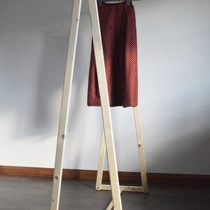 May include: A wooden clothing rack with a skirt hanging on a hanger. The skirt is a dark red color with small white polka dots. The rack is made of light-colored wood and has an A-frame design.